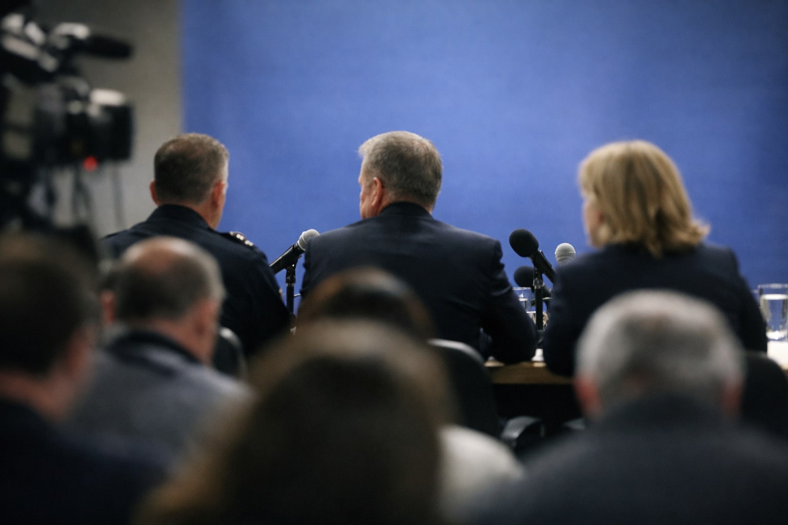 Suasana konferensi pers dengan pejabat duduk di meja konferensi dilihat dari belakang atau samping tanpa wajah terlihat jelas